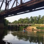 people in a blue kayak on the river surrounded on the banks with leafy trees and some sandy beachs