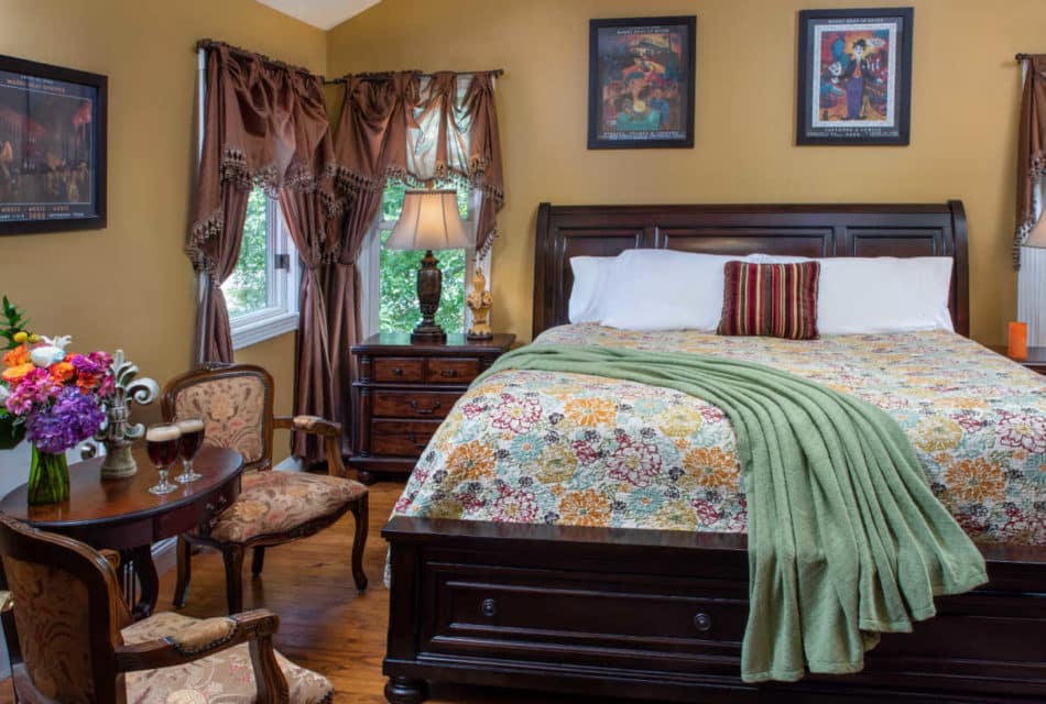 large high bed with yellow and green flowered spread in the front are 2 chairs next to a table