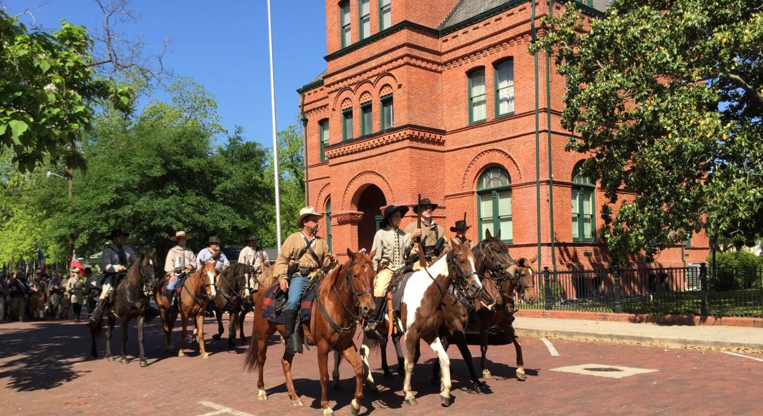 Men riding horses on a cobblestone road with large red brick building and green trees in the background